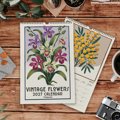 a vintage-style wall calendar with a floral design, placed on a wooden surface alongside other items such as a camera, a mug, and some plants.
