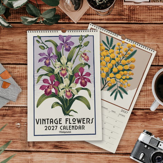 a vintage-style wall calendar with a floral design, placed on a wooden surface alongside other items such as a camera, a mug, and some plants.